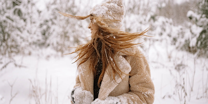 Woman in a snow-covered coat and beanie turns joyfully in a winter landscape—capturing the need for nourishing skincare during the cold Aussie season.