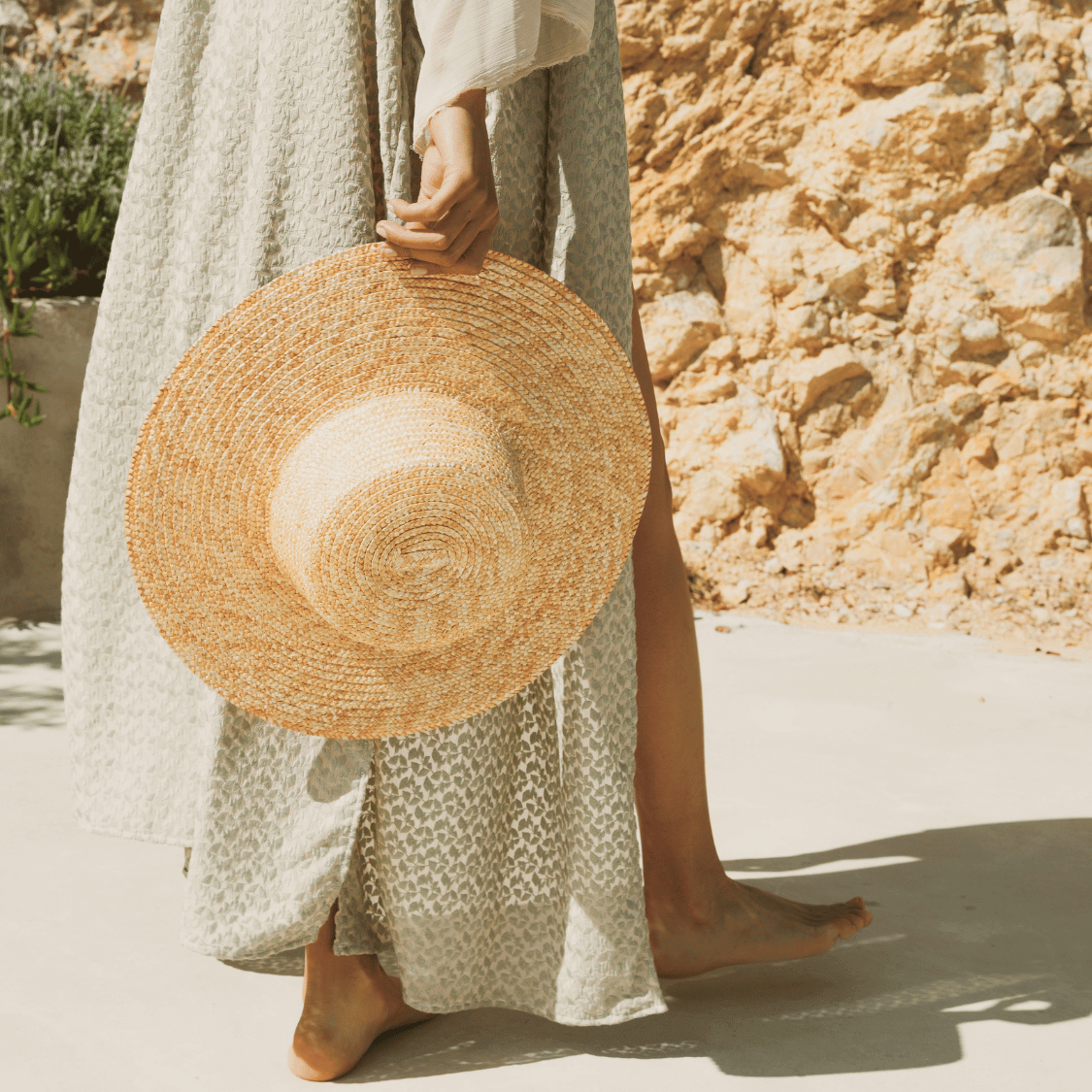 A barefoot woman in a flowy printed dress holds a straw sunhat by her side while standing on a sunlit stone path. The relaxed, summery scene evokes effortless holiday beauty—perfectly pairing with the Glow Kit Travel Size from Karin Herzog for radiant, on-the-go skincare.