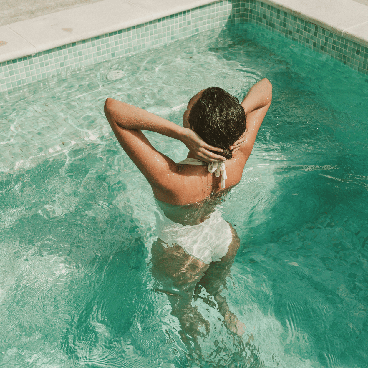A woman in a white swimsuit stands in a turquoise tiled pool with her back to the camera, gently holding her wet hair under the sun. The clear water ripples around her, creating a relaxed, summery atmosphere—an ideal moment for the Karin Herzog Glow Kit in Travel Size, perfect for glowing skin poolside.