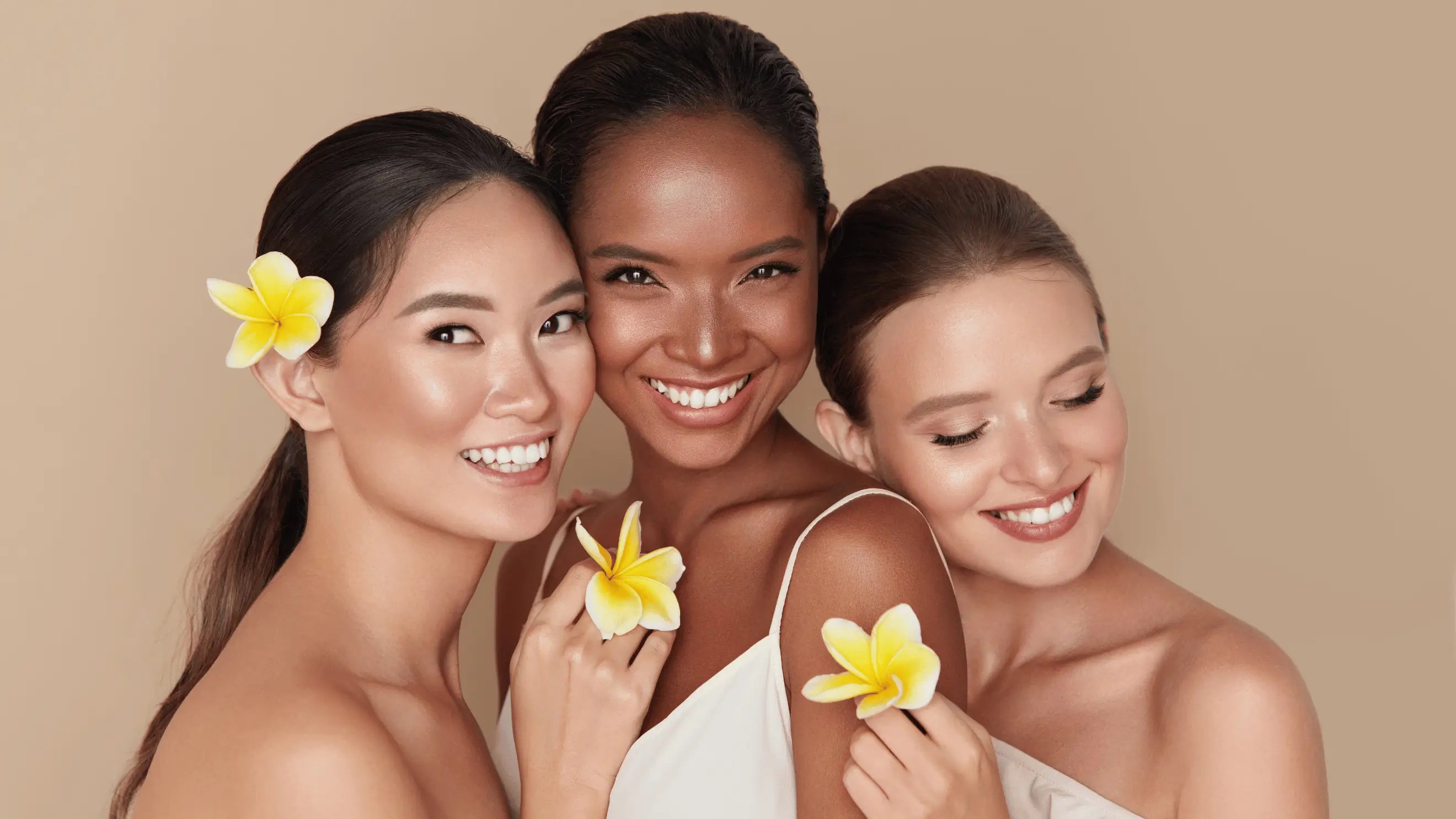 Three radiant women smiling together with fresh flowers, showcasing healthy, glowing skin — a celebration of beauty and friendship for Karin Herzog's Refer a Friend program.