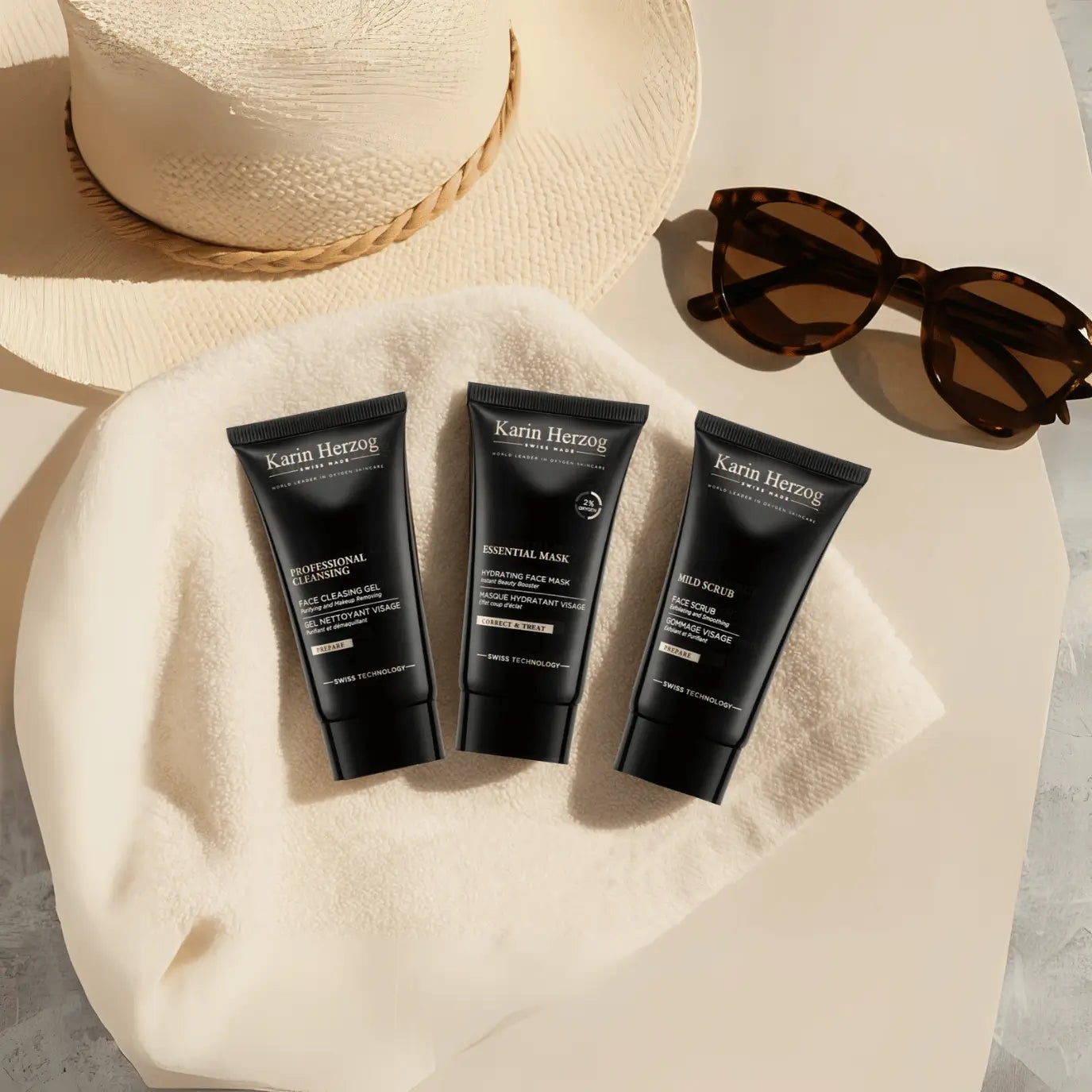 Flatlay image of the Karin Herzog Glow Kit – Travel Size, featuring three 25ml black skincare tubes (Professional Cleansing, Essential Mask, and Mild Scrub) arranged on a soft beige towel. A woven straw hat and tortoiseshell sunglasses complete the summery, poolside setting under natural sunlight.
