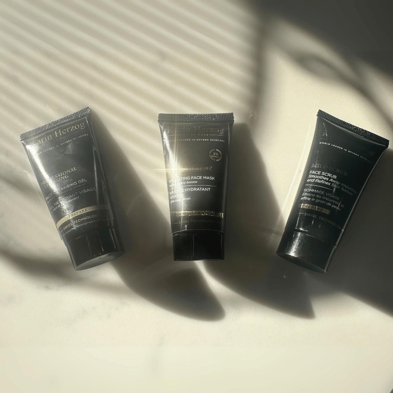 Three Karin Herzog Glow Kit 25ml skincare tubes—Professional Cleansing, Essential Mask, and Mild Scrub—are arranged on a smooth surface with soft natural sunlight casting diagonal shadows through window blinds, creating a warm and moody studio feel.