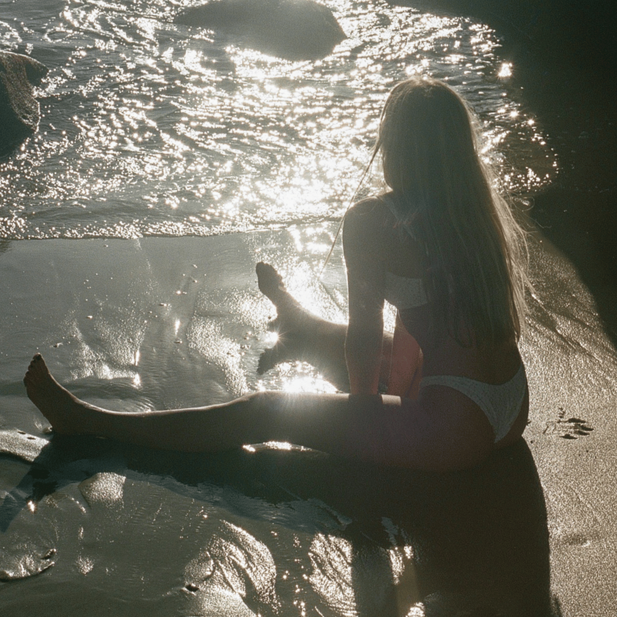 A woman in a white bikini sits at the edge of the ocean, her body glowing in the soft, golden light of sunset. The gentle waves reflect the sun, creating a serene and radiant beachside moment—enhanced by the nourishing effects of the Karin Herzog Glow Kit in Travel Size.