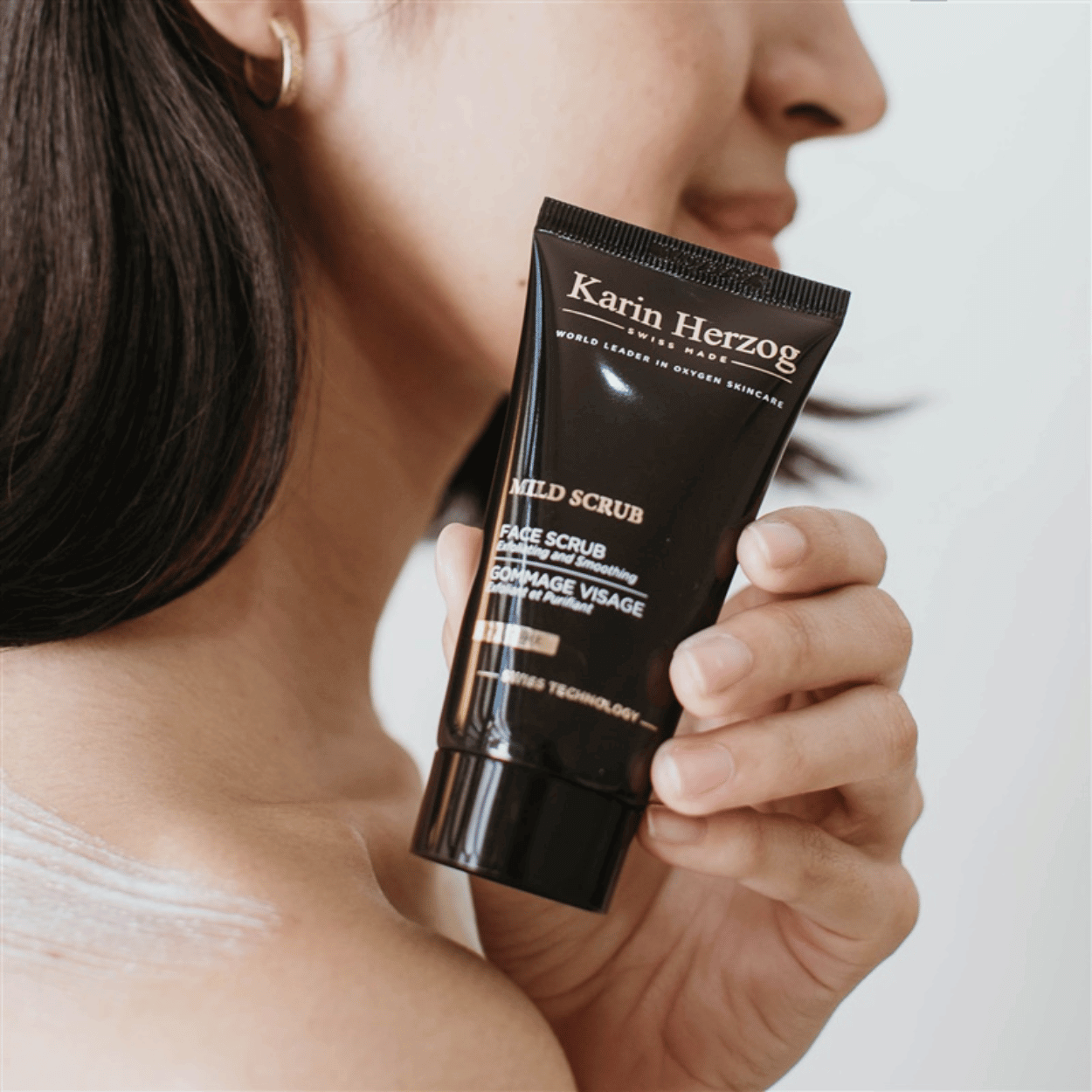 A woman holding a bottle of Karin Herzog Mild Scrub, showcasing the skincare product.