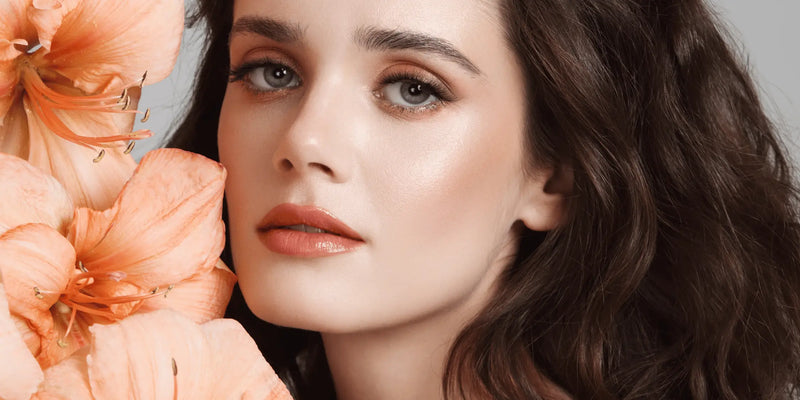 Close-up of a woman with radiant skin, soft coral makeup, and voluminous brown hair, surrounded by peach flowers — showcasing a glowing look inspired by Karin Herzog’s Beautify Makeup.