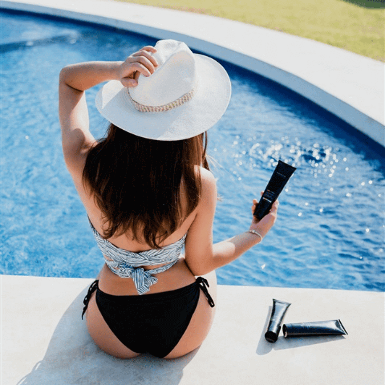 A woman in a black bikini and sunhat sits poolside, holding a tube of Karin Herzog Tone & Tan. With glowing skin and summer-ready style, she prepares to apply the self-tanning cream for a radiant, even tan. Two additional product tubes lie beside her on the pool ledge.