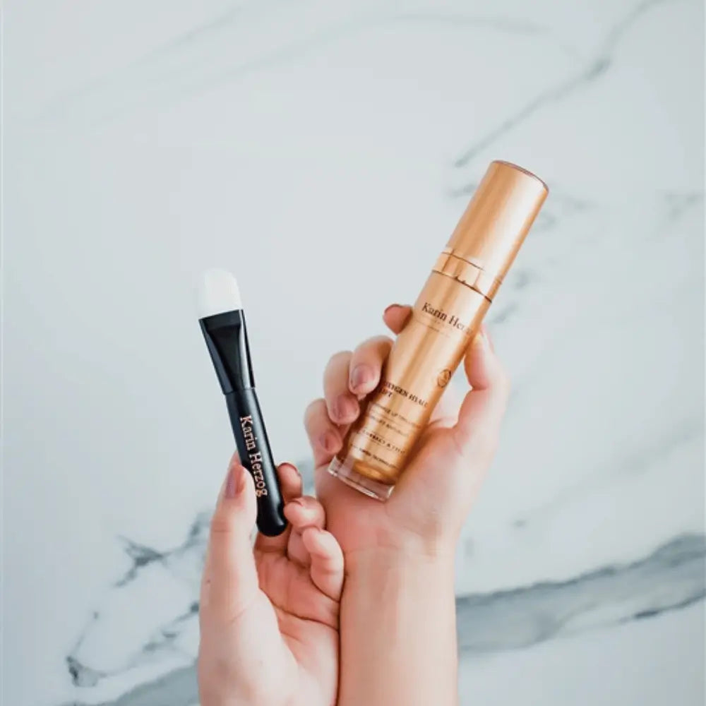 Hands holding the Karin Herzog Oxygen Hyalu Lift 30ml gold bottle alongside a precision application brush, set against a soft marble background.