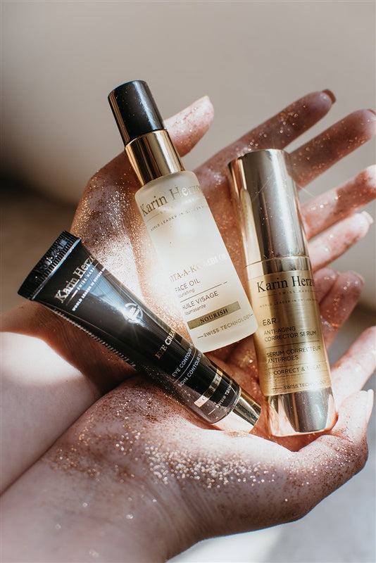 A woman’s glitter-adorned hand holding Karin Herzog Eye Cream, Vita-Kombi Oil, and E&R Anti-Ageing Corrector Serum, showcasing premium skincare products.