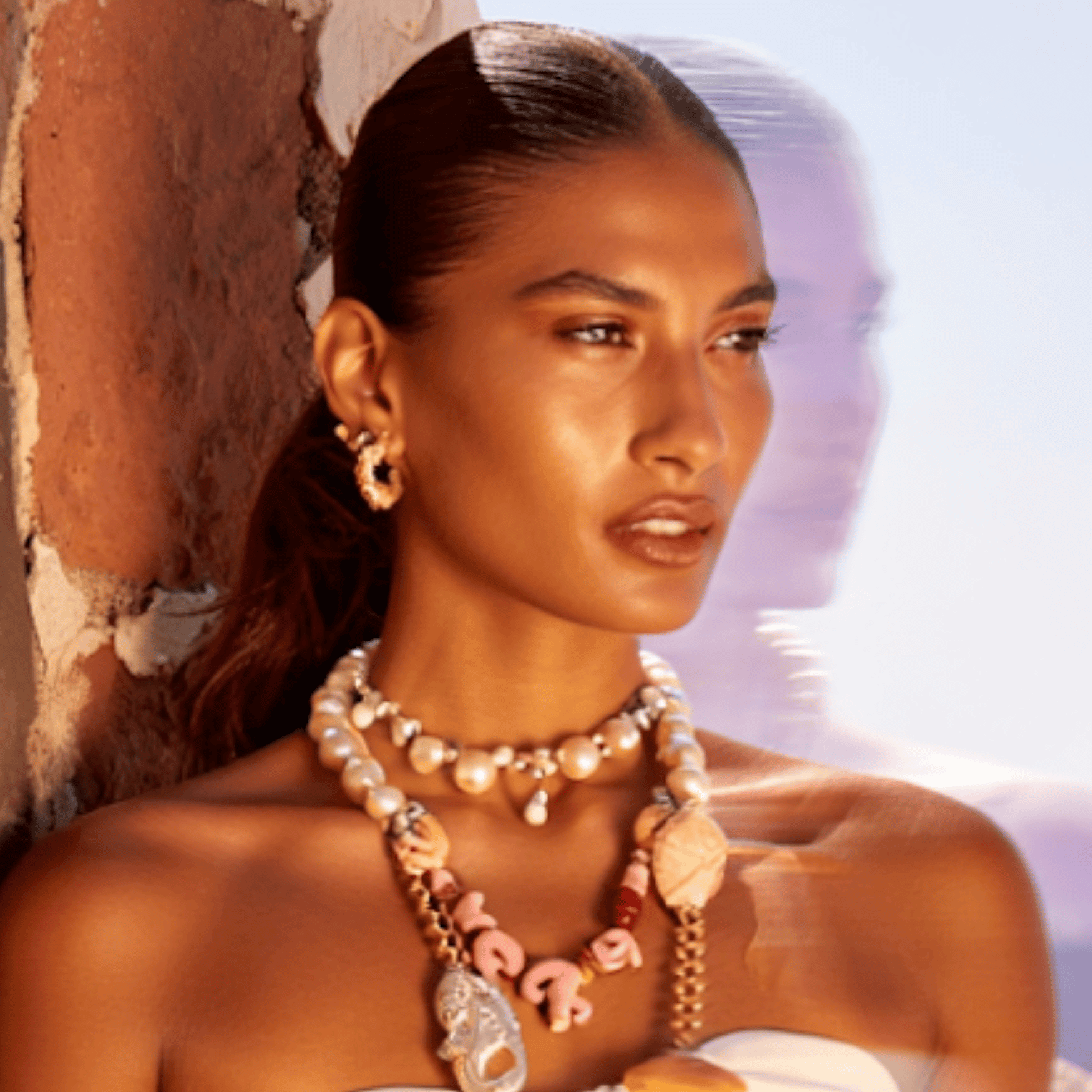 Sun-kissed model with glowing, radiant skin wearing bold pearl and shell jewellery, embodying the luminous effect of Karin Herzog’s The Golden Hour skincare.