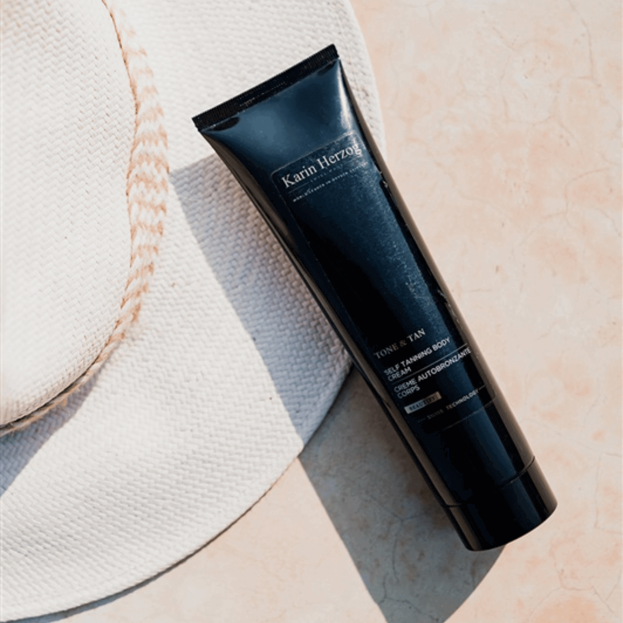 Flatlay of Karin Herzog Tone & Tan self-tanning body cream placed beside a white sunhat on a peach-toned surface, highlighting the product’s sleek black packaging and summer-ready appeal.